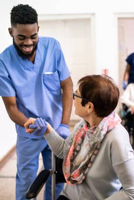 A smiling male healthcare workers assists an elderly female patient in her wheelchair, in the background another healthcare worker pushes a male patient